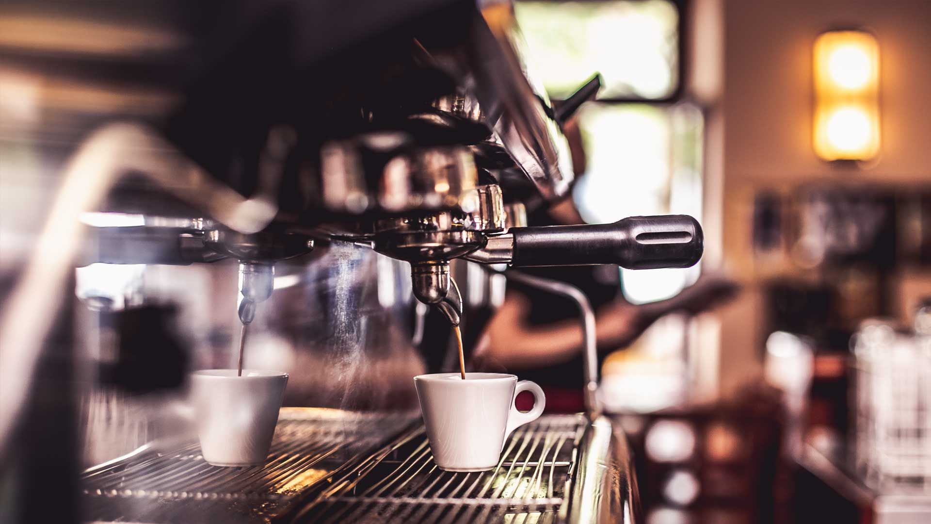 Siebträgermaschine in einem Café: Espresso läuft in zwei weiße Tassen, im Hintergrund warmes Licht.