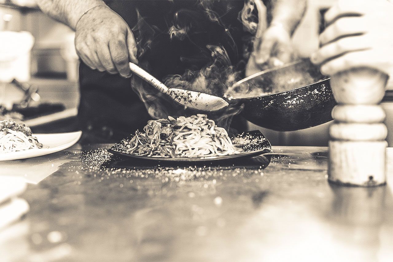 Schwarz-Weiß-Foto aus einer Restaurantküche: Ein Koch gibt dampfende Pasta aus der Pfanne auf einen dunklen Teller, daneben eine Pfeffermühle.