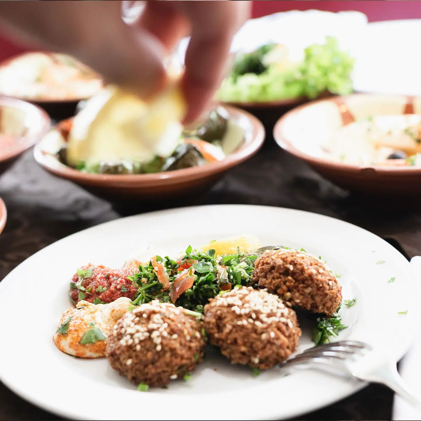 Teller mit Falafel, Taboulé, Hummus und Muhammara, im Hintergrund weitere Schalen; eine Hand träufelt Zitrone über den Salat.